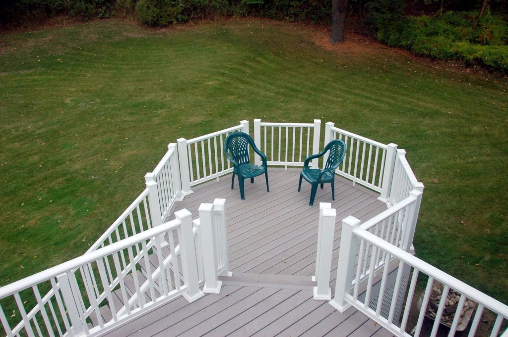 A high-angle view of a grey deck with white railings, two green chairs, and a stairway overlooking a grassy lawn.
