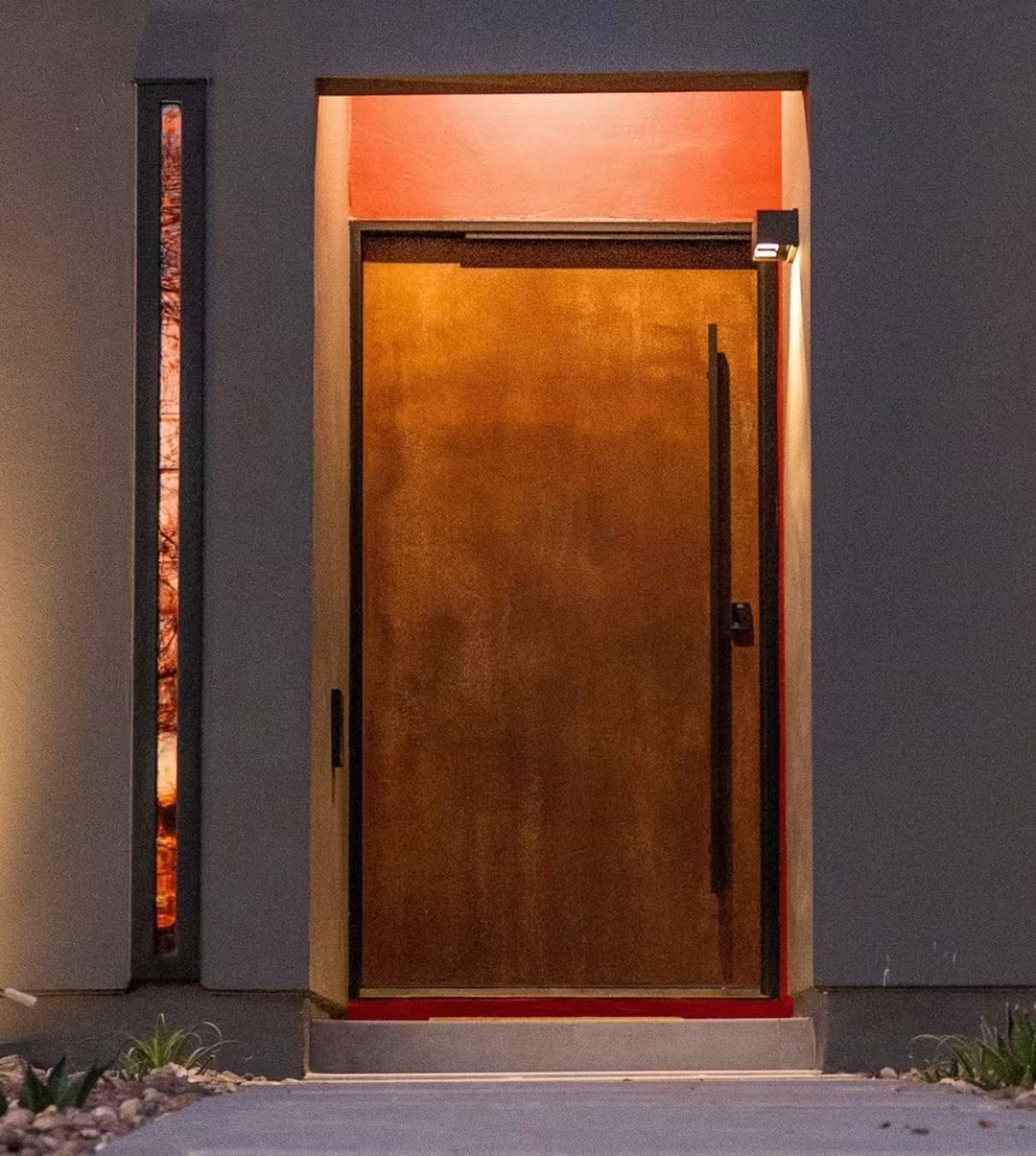 Modern rust-finish pivot entry door with a tall pull handle, lit at dusk